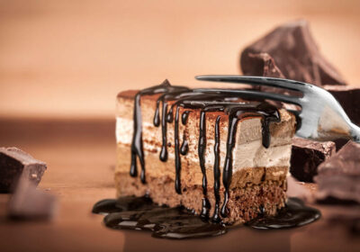 Chocolate layered dessert with drizzled chocolate sauce and a fork, surrounded by chocolate pieces, emphasizing indulgence in sweet treats.