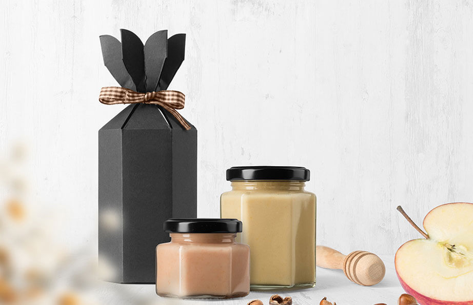 Black gift box with bow, two jars of creamy spreads, honey dipper, and sliced apple on a light wooden background, representing lifestyle and food themes.