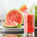 Watermelon smoothie in a glass with a slice of watermelon and cut watermelon pieces in the background, emphasizing refreshing summer drinks and healthy lifestyle choices.