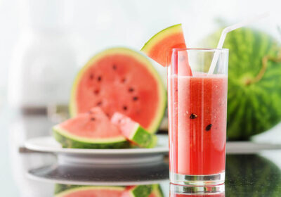 Watermelon juice in a tall glass with a slice of watermelon garnish, fresh watermelon halves in the background, highlighting refreshing summer beverages.