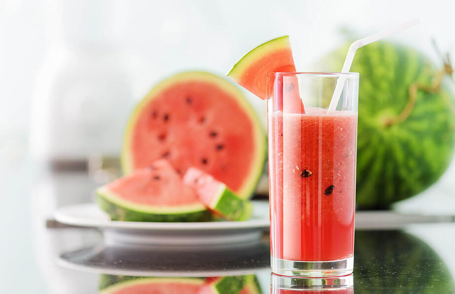 Fresh watermelon juice in a tall glass garnished with a slice of watermelon, with cut watermelon pieces and whole watermelons in the background, highlighting summer refreshment and healthy lifestyle.