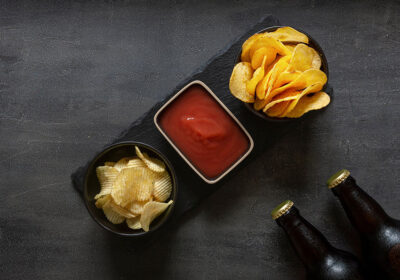 Chips with ketchup served on a slate platter alongside two bottles of dark beer, highlighting snack options for social gatherings.