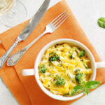 Bowl of baked pasta with broccoli and cheese on an orange cloth, accompanied by a fork and knife, highlighting a delicious hobby-related dish.