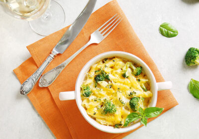 Bowl of creamy pasta with broccoli, garnished with fresh basil, on an orange napkin, accompanied by a glass of white wine and silver cutlery.